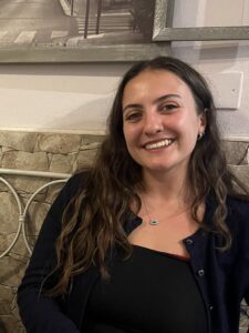 Megan Ioannides, a womena with long brown hair and wearing a black top and jacket, smiles to camera.