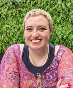 Minna, who has short blonde hair, smiles to camera. They are wearing a pink and purple patterned top, in front of a green backdrop of plants