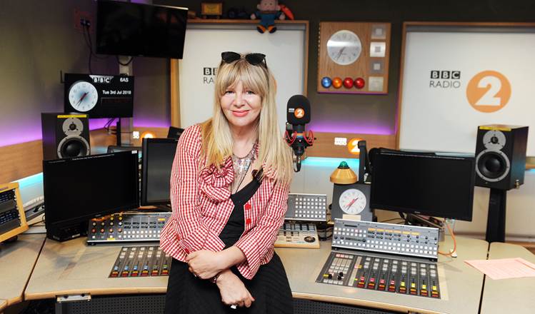 Janey Lee Grace is a blonde woman wearing a red check shirt. She sits in a radio production studio, smiling.