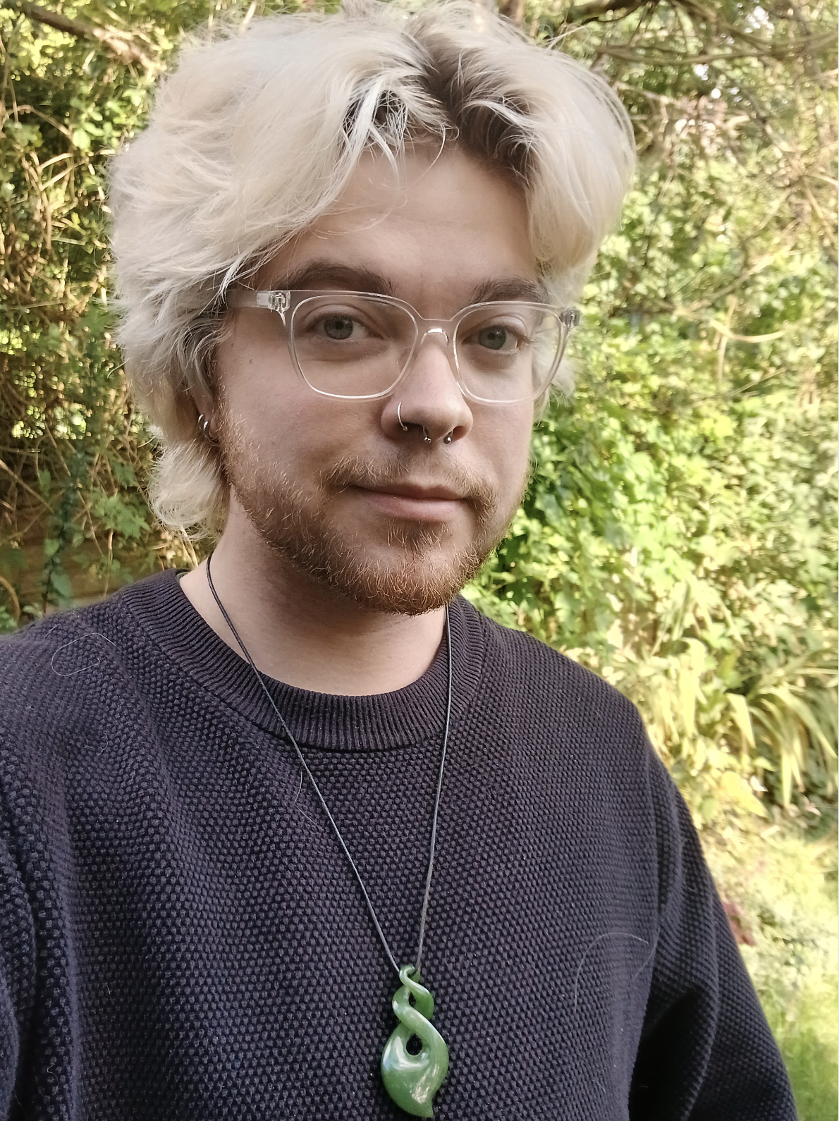 Alex, a blonde haired man with clear glasses, stands in front of greenery