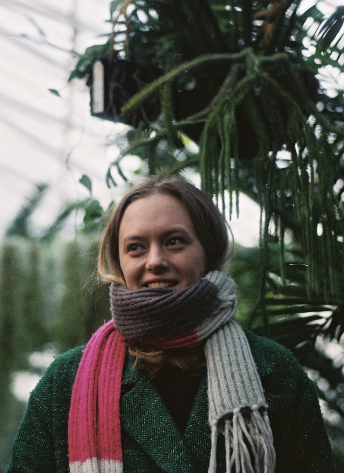 Lucy wears a green jacket and a colourful knitted scarf. In the background, a greenhouse and plants can be seen in soft focus.