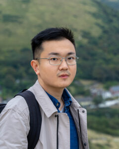 A person with short black hair, glasses, a light mustache, and a neutral expression looking directly at the camera, wearing a beige jacket and blue shirt with a black backpack. The background is outdoors with soft focus greenery and hills.