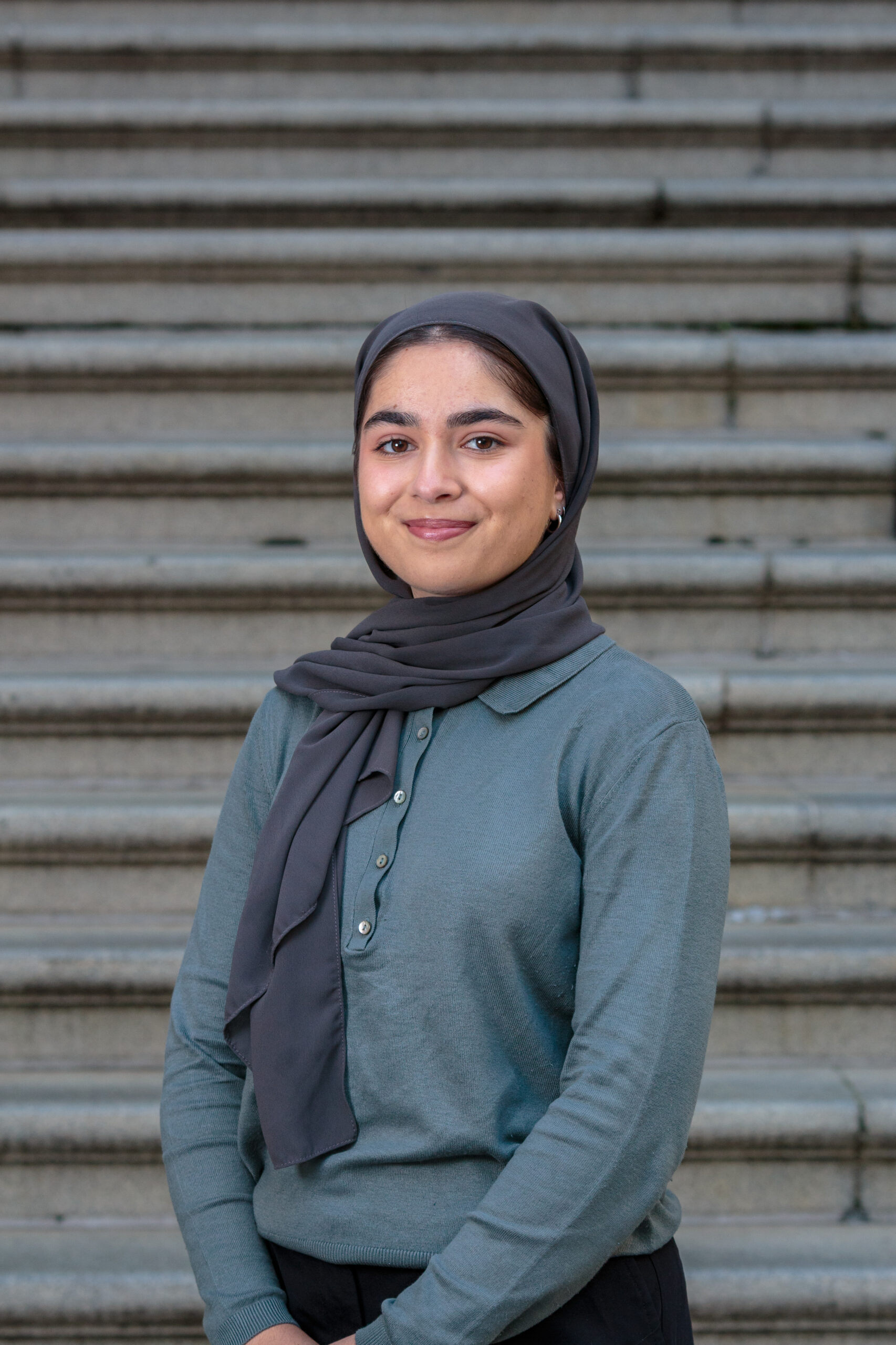 sahar, a woman wearing a grey green top and a black headscarf, stands infront of grey steps