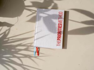 The Priorities Method Journal – a cream bound book with red lettering – on a white tabletop.