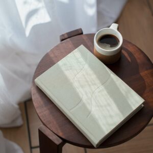 The Give Yourself Kindness Journal – a sage coloured bound book – rest on a round bedside table next to a coffee.